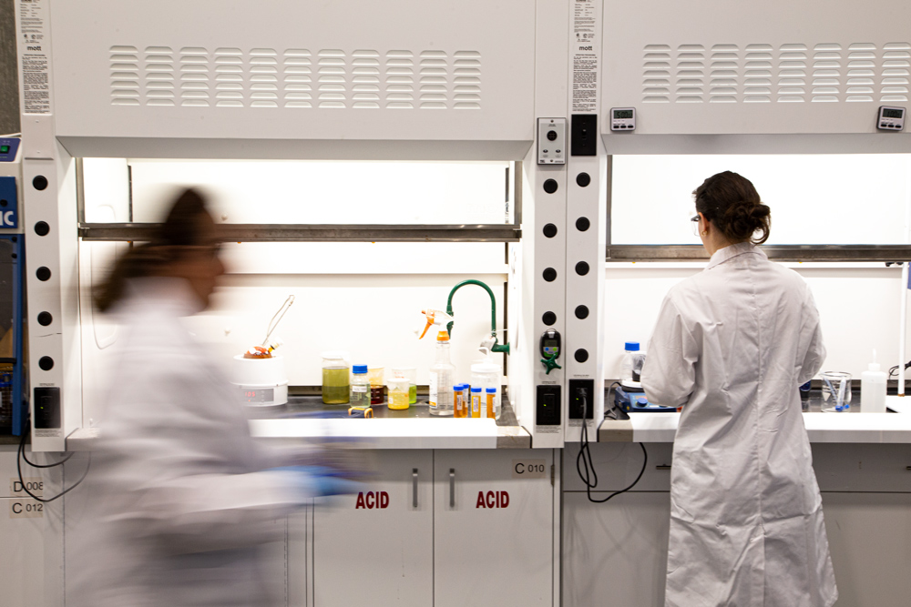 Two scientists in a laboratory; one in focus working at a fume hood with various chemicals, while the other moves quickly in the foreground, creating a blurred motion effect.